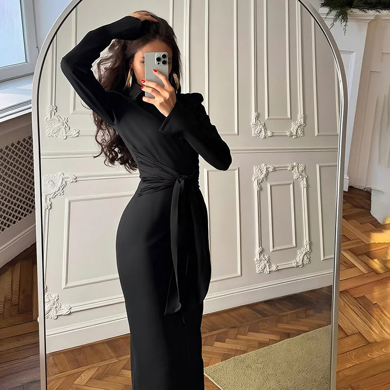 Person taking a mirror selfie wearing a black dress in a room with wooden flooring and white walls.