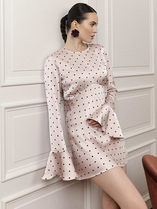 Woman wearing a polka dot dress with bell sleeves against a white paneled wall.