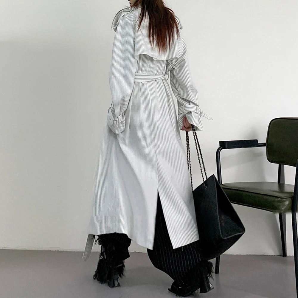 Person wearing a long, light-colored coat with a black chair in the background