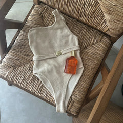 Beige one-piece swimsuit with belt and tanning oil on a chair