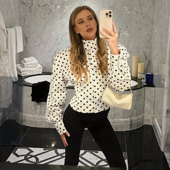 Woman taking a mirror selfie in a bathroom wearing a polka dot blouse and black pants.