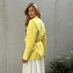 Woman wearing a yellow jacket and white skirt against a gray wall with plants.