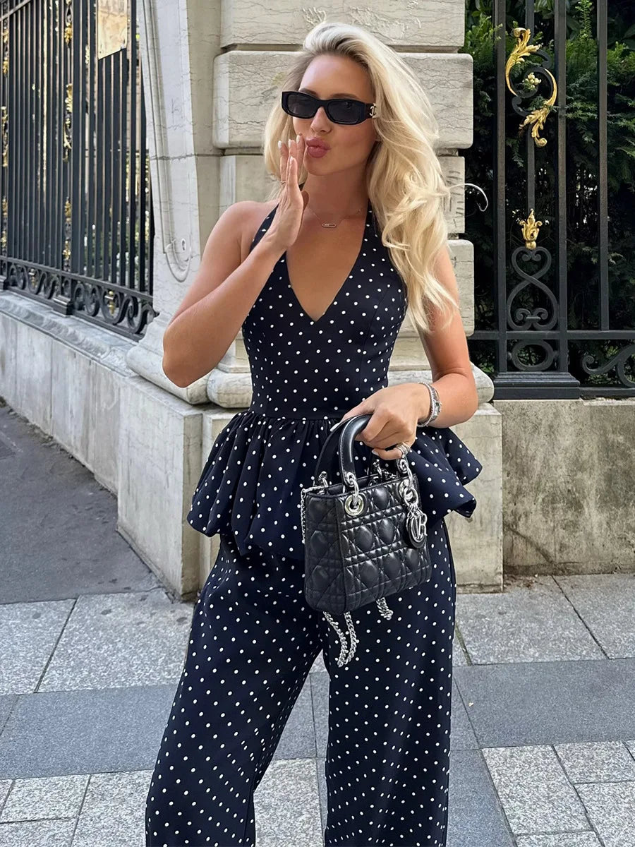 Woman in a black polka dot jumpsuit with sunglasses and a handbag, standing on a sidewalk.