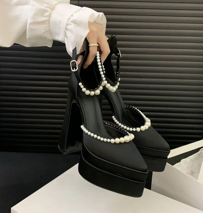 Black high-heeled shoes with pearl straps held by a hand against a neutral background
