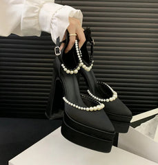 Black high-heeled shoes with pearl straps held by a hand against a neutral background