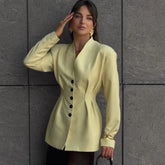 Woman wearing a light green blazer with black buttons against a gray tiled wall.