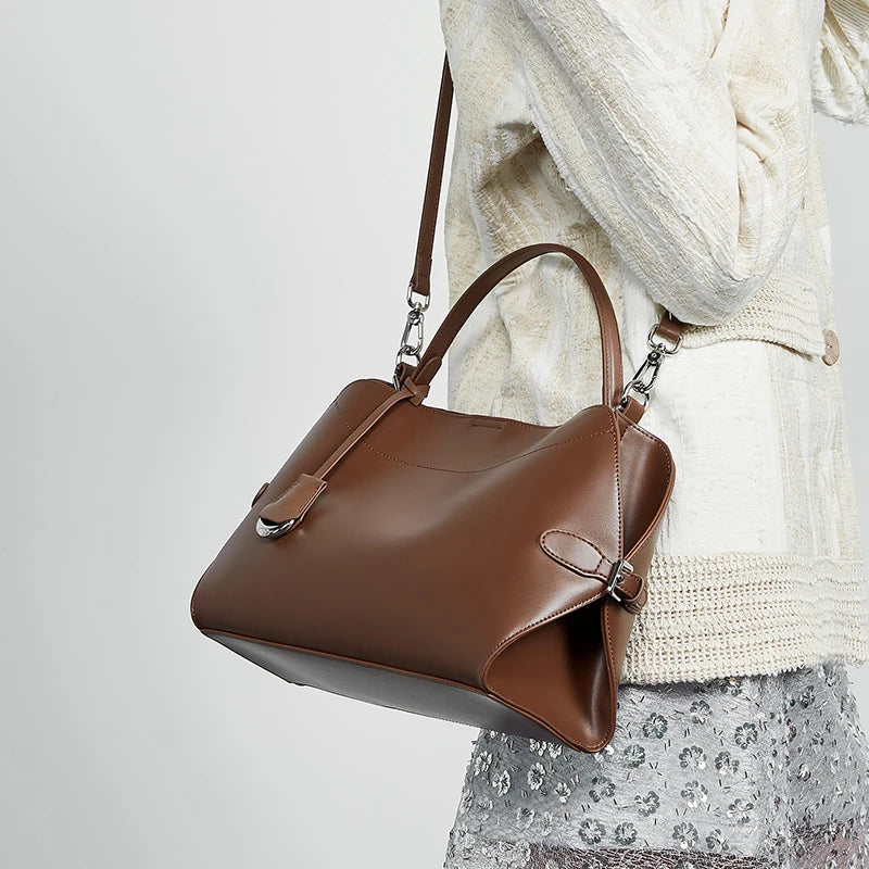 Brown handbag held by a person wearing a white sweater and patterned skirt on a light gray background