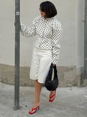 Woman wearing a polka dot blouse, white shorts, red shoes, and holding a black bag against a plain wall.