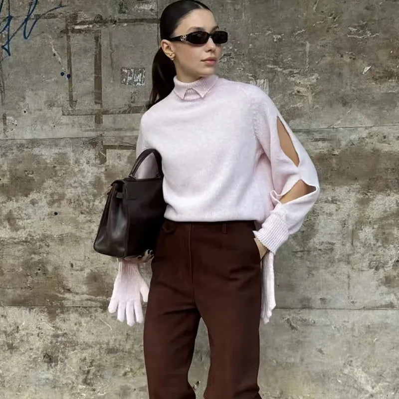 Woman wearing a light pink sweater with cut-out shoulders against a textured wall.