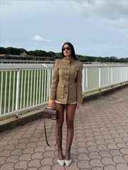 Woman in a beige military-style jacket standing on a paved walkway with a railing and greenery in the background.