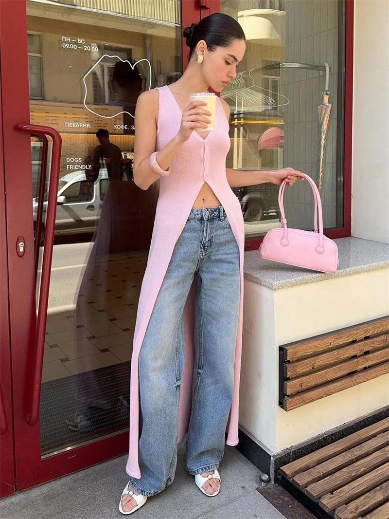 Woman in pink cardigan and blue jeans holding a pink handbag and drink, standing in front of a store.