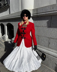 Woman in a red coat and white dress standing against a stone wall.
