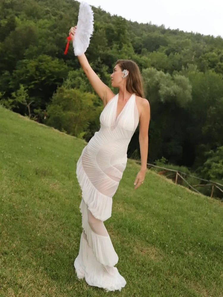 Woman in a white dress holding a white feathered object on a grassy hill with trees in the background