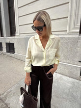 Woman wearing a light-colored blouse with a logo and dark pants, standing on a street.