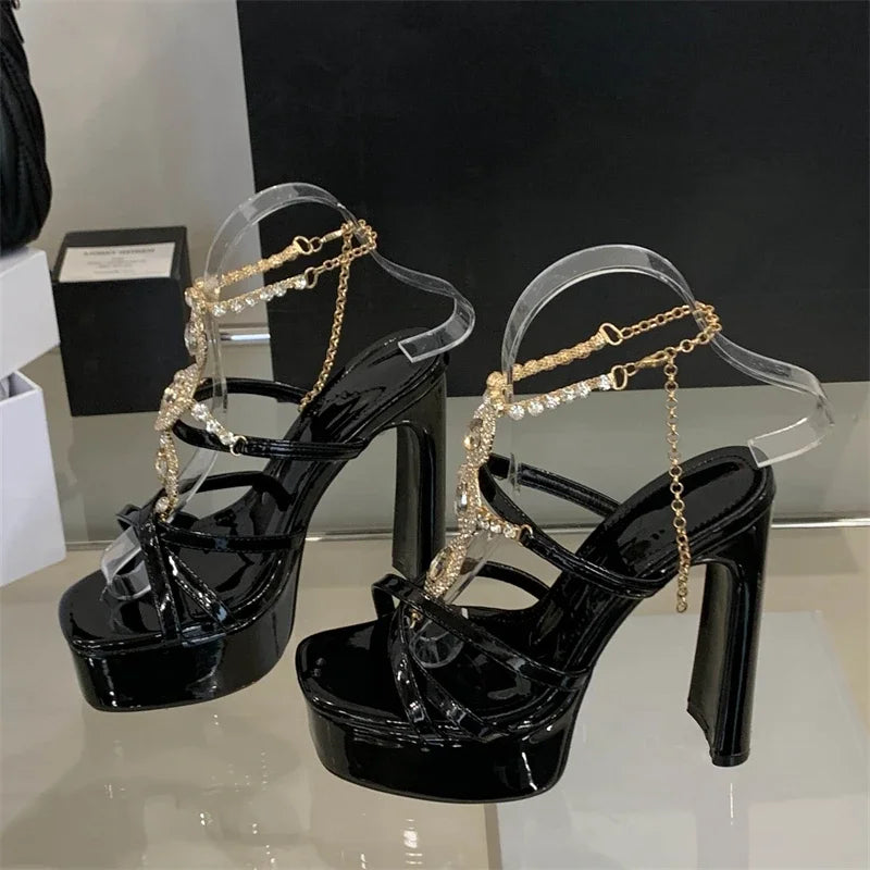 Black high-heeled shoes with gold chain straps on a reflective surface.