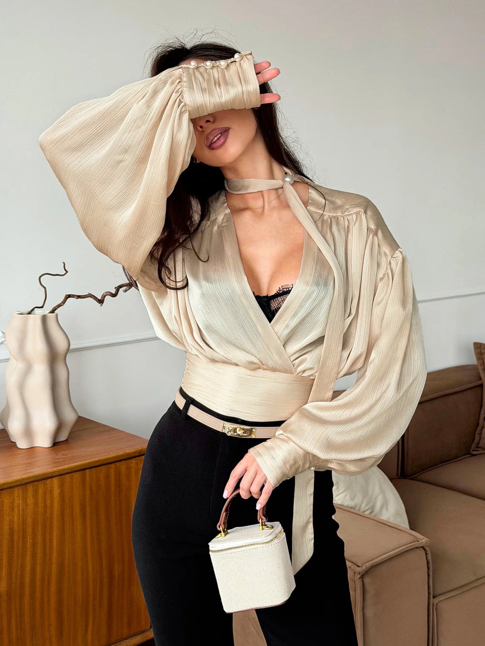 Woman in a beige blouse and black pants holding a white handbag in a living room setting.