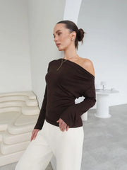 Woman wearing a brown one-shoulder top and white pants in a minimalistic room.