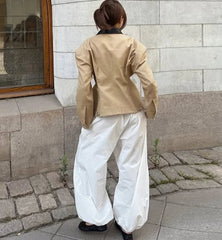 Person wearing a beige jacket and white pants standing against a stone wall.