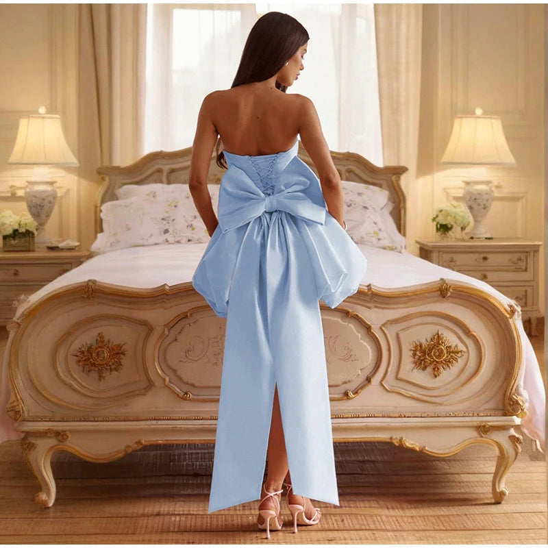 Woman in a light blue dress with a large bow standing in a bedroom.