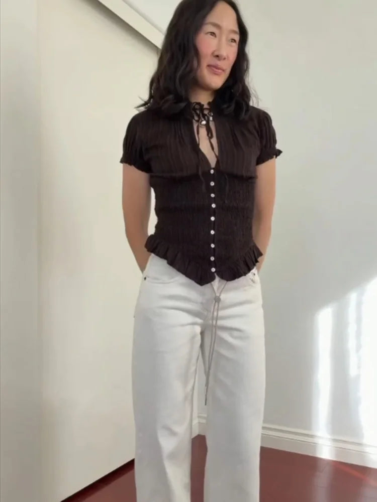 Woman wearing a brown top with ruffled details and white pants in a room with a white wall.