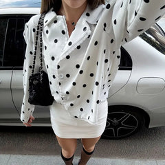 Person wearing a white polka dot blouse and skirt with a black handbag in front of a white car.