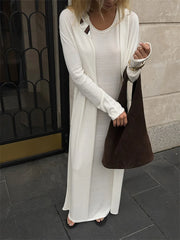 Woman wearing a long white cardigan over a white dress, holding a brown bag on a city street.