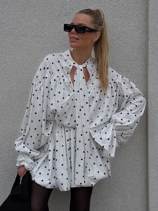 Woman wearing a white blouse with black polka dots against a gray wall.