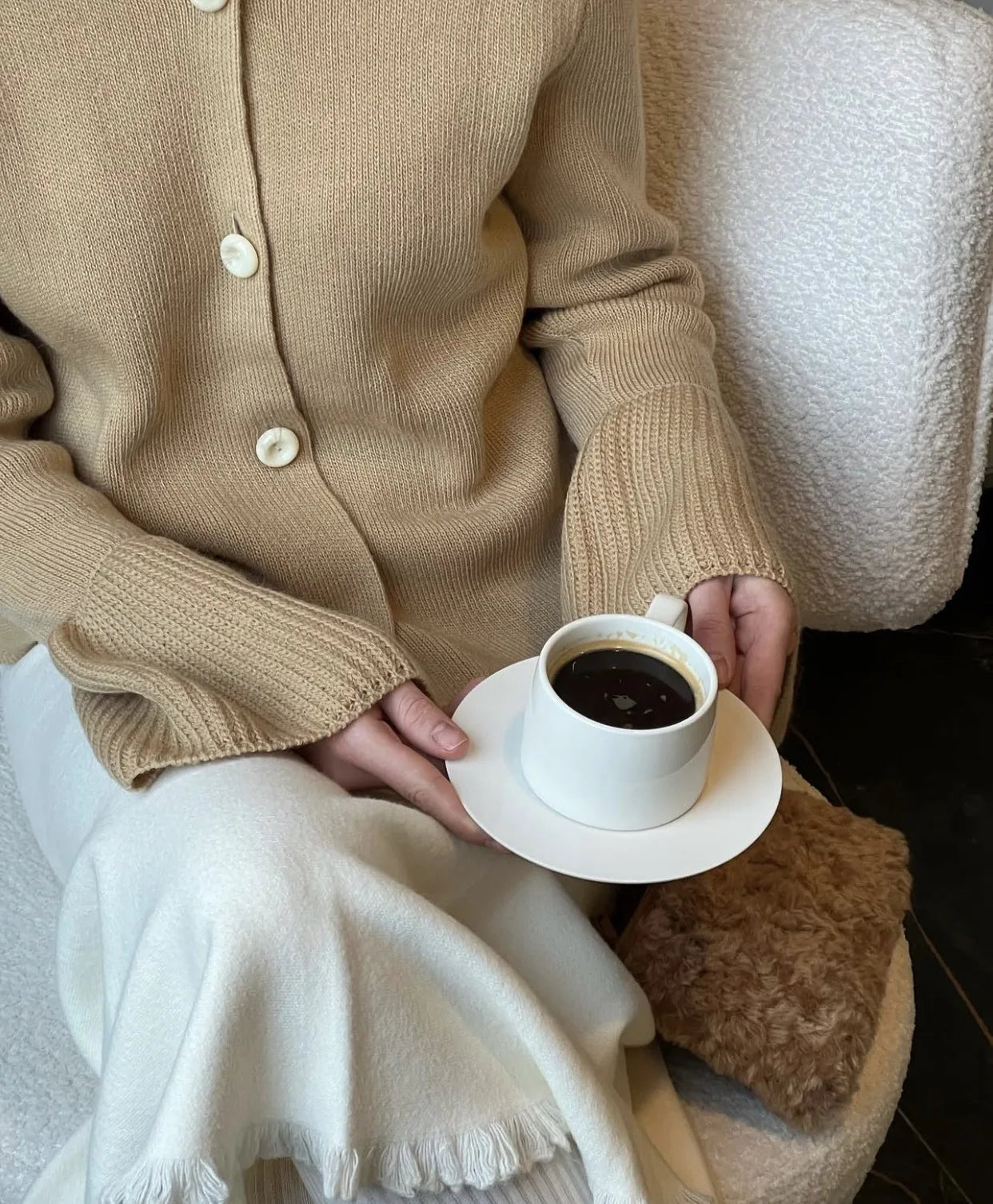 Person wearing a beige cardigan holding a white cup of coffee on a saucer.