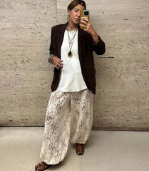 Person taking a mirror selfie wearing a white top, brown blazer, and patterned pants against a textured wall.
