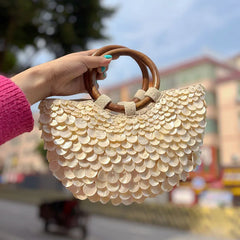 Hand holding a shell-patterned handbag with wooden handles against an outdoor background
