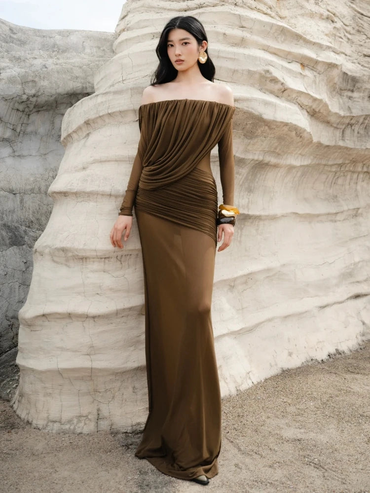 Woman in a brown off-shoulder dress standing against a textured white wall.