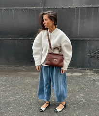 Woman wearing a white sweater, blue jeans, and carrying a brown bag against a dark wall.