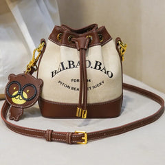 Beige canvas bucket bag with brown faux leather trim and bear charm.