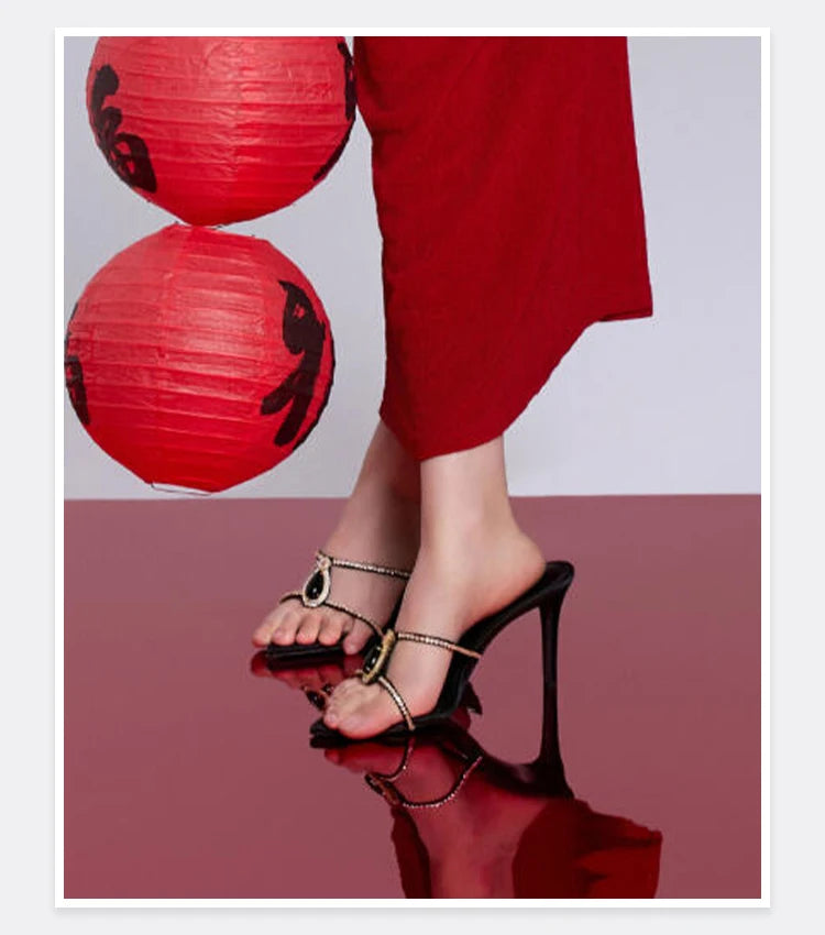 Black high-heeled sandals worn with a red outfit against a reflective surface.
