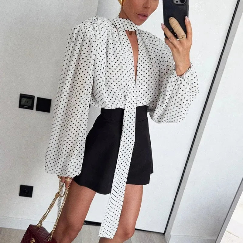 Woman taking a mirror selfie wearing a white blouse with black polka dots and black shorts.