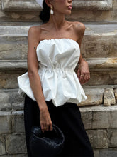 Woman wearing a white strapless top and black pants against a stone wall.