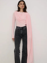 Woman wearing a pink long-sleeve top and black jeans with a pink scarf draped over her shoulders against a white background.