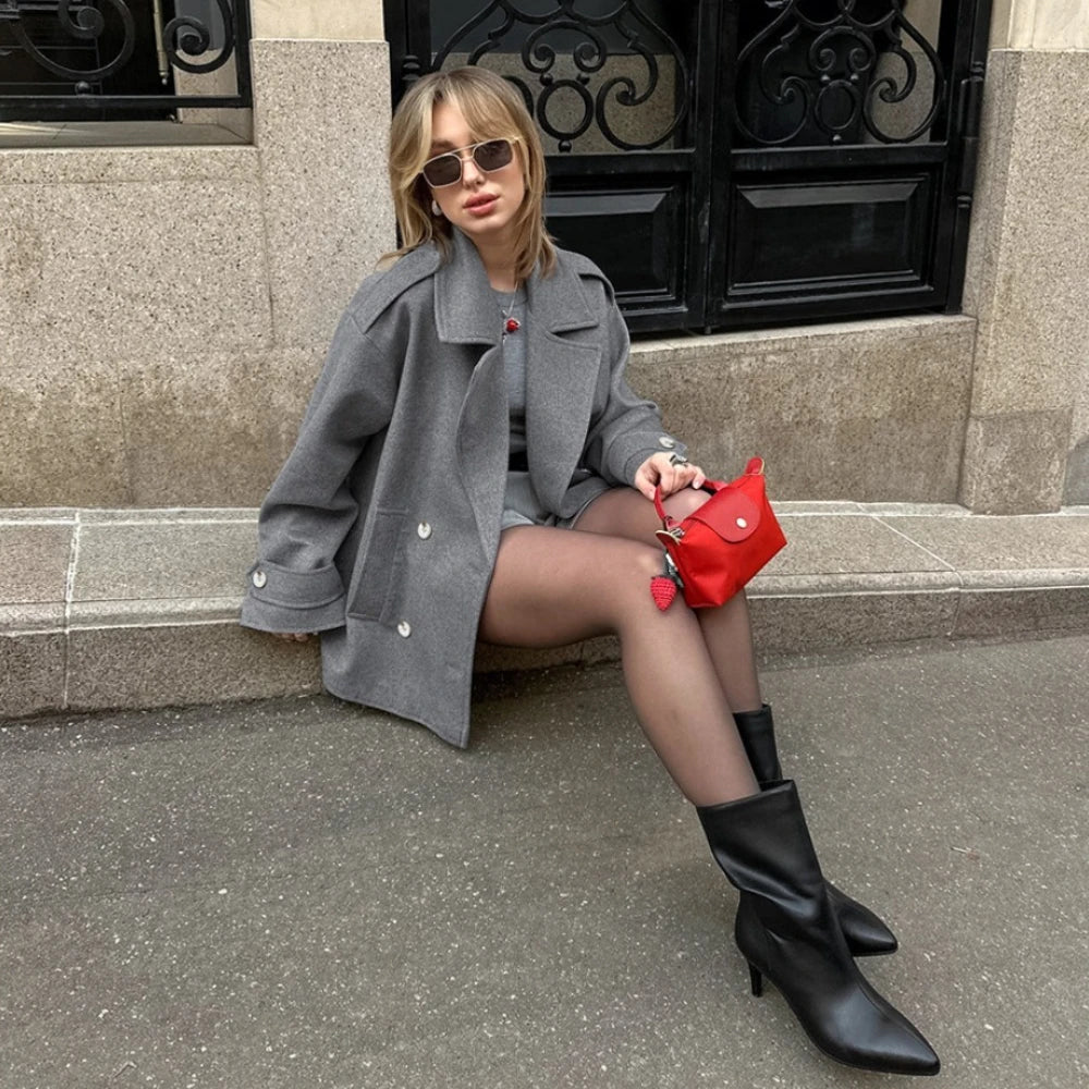 Woman in gray coat and black boots sitting on steps with a red handbag.