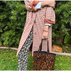 Person wearing a red and white checkered coat with leopard print pants and bag, standing in front of green foliage.