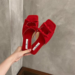 Red velvet high-heeled shoes with bow detail held by a hand against a gray wall.