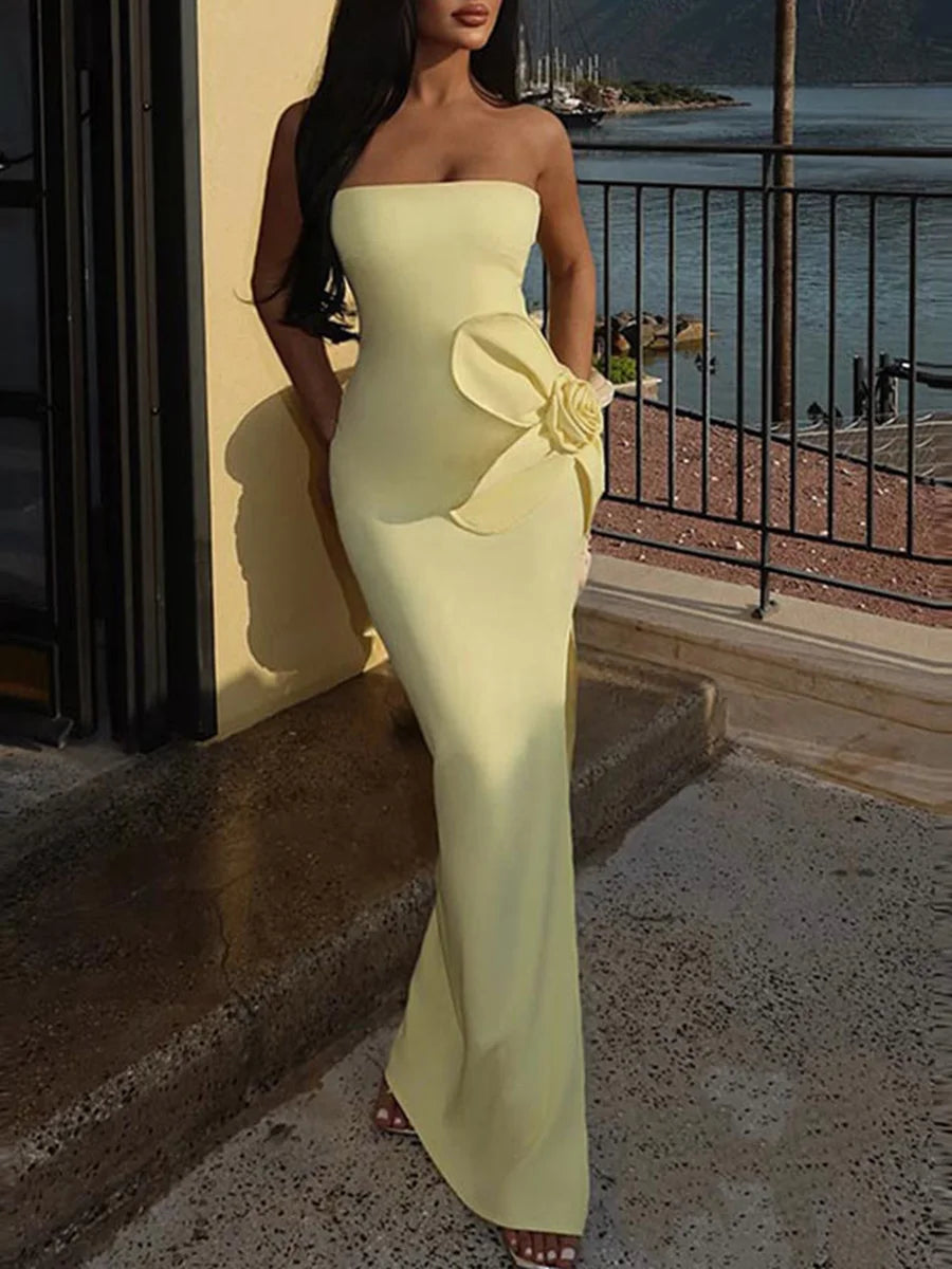 Woman in a strapless yellow evening gown with a large flower detail, standing on a balcony with a waterfront view.