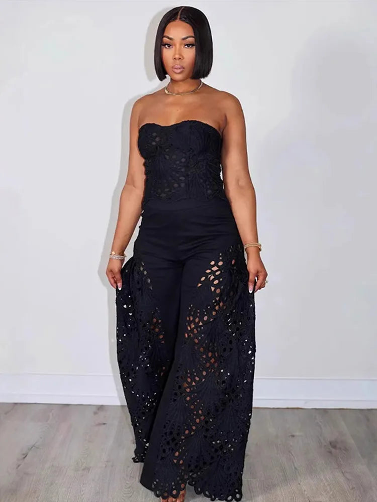 Woman wearing a black strapless top and wide-leg pants with cut-out details against a white wall.