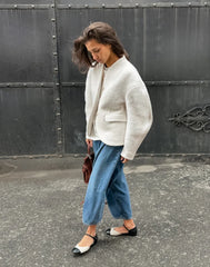 Woman wearing a white jacket and blue pants standing against a dark metal gate.