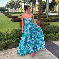 Woman in a blue floral dress standing in a garden with palm trees and greenery.