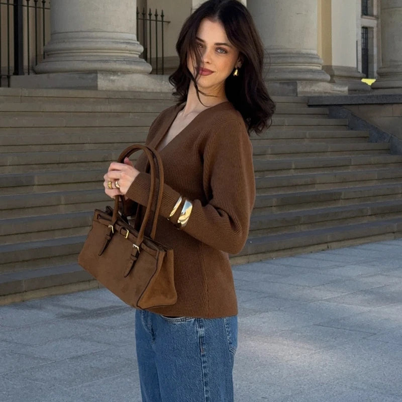 Woman in a brown sweater and blue jeans holding a brown handbag on stone steps.