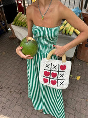 Woman in a green and white striped dress holding a coconuts and a woven bag with a tic-tac-toe pattern.