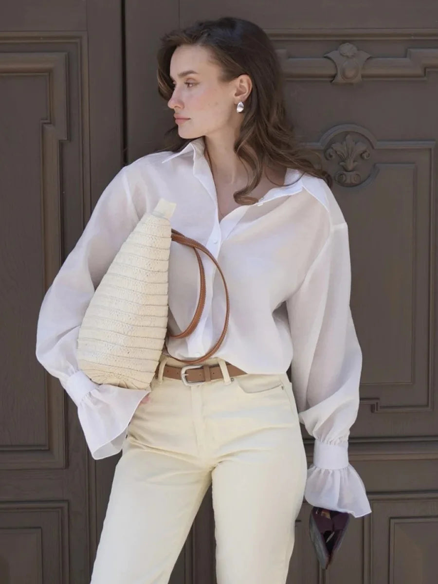 Woman wearing a white blouse and beige pants, holding a woven bag, standing in front of a wooden door.