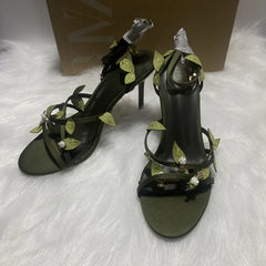 Black high-heeled sandals with green leaf embellishments on a white surface.