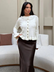 Woman wearing a white jacket with a ruffled collar and brown skirt in a living room setting.
