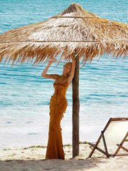 Woman in a yellow dress standing under a thatched beach umbrella with clear blue water and sand in the background.